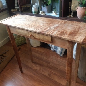 May include: A wooden console table with a single drawer and a natural wood finish. The table has a unique wood grain pattern and is supported by four legs.