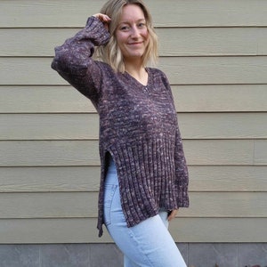 May include: A woman wearing a purple and pink knitted sweater with side slits and light blue jeans.