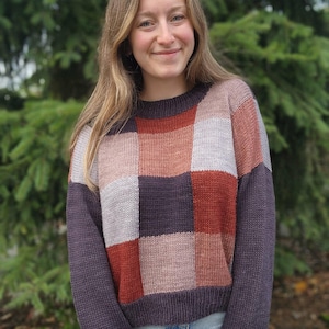 May include: A patchwork sweater in shades of purple, rust, pink, and gray. The sweater has a crew neck and long sleeves. The model is wearing blue jeans. The background is a blurred green tree.