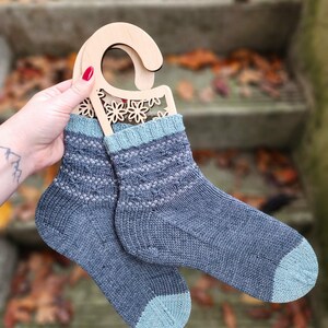 May include: A pair of knitted socks in shades of blue and teal, displayed on a wooden hanger. The socks feature a patterned design on the upper portion and solid teal toes and cuffs. The wooden hanger has a floral cutout design.