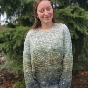 May include: A crew neck sweater with a gradient color scheme, transitioning from light blue to green and ending in a dark blue at the cuffs. The sweater is made of a knitted material and is worn with blue jeans.