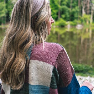 Op de afbeelding: Een vrouw draagt een lichtroze geknoopte hoofdband en een patchworktrui. De trui heeft vierkanten in wit, blauw, groen en kastanjebruin. De achtergrond toont een meer en bomen.