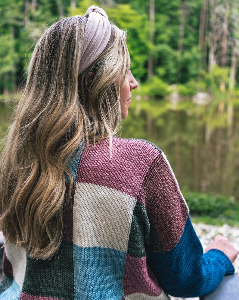 Op de afbeelding: Een vrouw draagt een lichtroze geknoopte hoofdband en een patchworktrui. De trui heeft vierkanten in wit, blauw, groen en kastanjebruin. De achtergrond toont een meer en bomen.