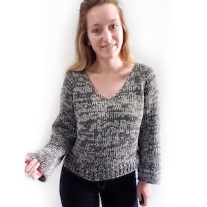 Puede incluir: Un suéter de punto gris y blanco con cuello en V y mangas largas. El suéter tiene un corte holgado y una longitud ligeramente recortada.