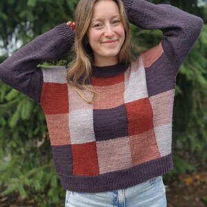 Op de afbeelding: Een gebreide patchworktrui in tinten paars, roest en grijs. De trui heeft een dambordpatroon over de borst en mouwen. Het model draagt een lichtblauwe spijkerbroek.
