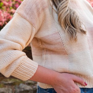 Op de afbeelding: Een cr&egrave;mekleurige gebreide trui met horizontale strepen in lichtroze en perzik. De trui heeft een geribbelde zoom en manchetten en een col. De mouwen zijn lang en de algehele stijl is casual en comfortabel.