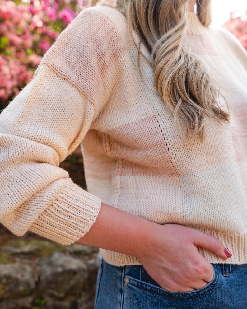 Op de afbeelding: Een cr&egrave;mekleurige gebreide trui met horizontale strepen in lichtroze en perzik. De trui heeft een geribbelde zoom en manchetten en een col. De mouwen zijn lang en de algehele stijl is casual en comfortabel.