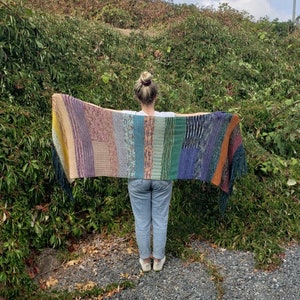 Pode incluir: Uma pessoa usando uma echarpe de malha listrada multicolorida com franjas. A echarpe é feita de diferentes tons de roxo, azul, verde, amarelo e laranja.