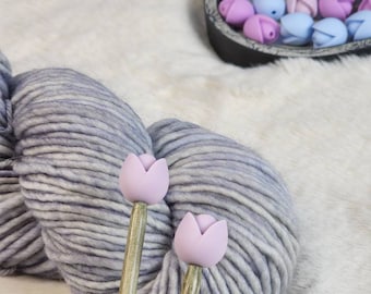 Puede incluir: Dos agujas de tejer de madera con puntas en forma de tulipán morado, junto a una bola de hilo gris.