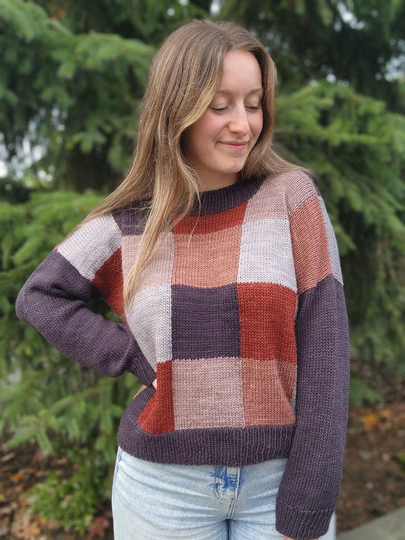 Op de afbeelding: Een patchwork trui in tinten paars, grijs en roest. De trui heeft een ronde hals en lange mouwen. Het ontwerp is een reeks vierkanten in verschillende kleuren. De mouwen en de halslijn zijn effen donkerpaars.