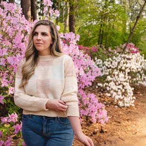 Op de afbeelding: Een cr&egrave;mekleurige trui met horizontale strepen in lichtroze en wit wordt gedragen met een blauwe spijkerbroek. De trui heeft een ronde halslijn en lange mouwen. De achtergrond toont roze en witte bloemen en bomen.