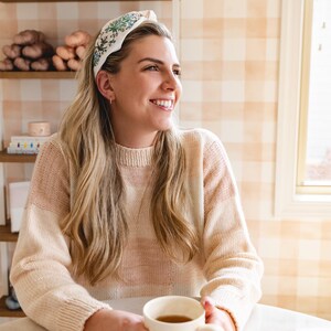 Op de afbeelding: Een cr&egrave;mekleurige hoofdband met een bloemenpatroon wordt gedragen door een persoon met lang blond haar. De persoon draagt een cr&egrave;me en roze trui en houdt een witte mok vast. De achtergrond heeft planken met garen en een geruite muur.