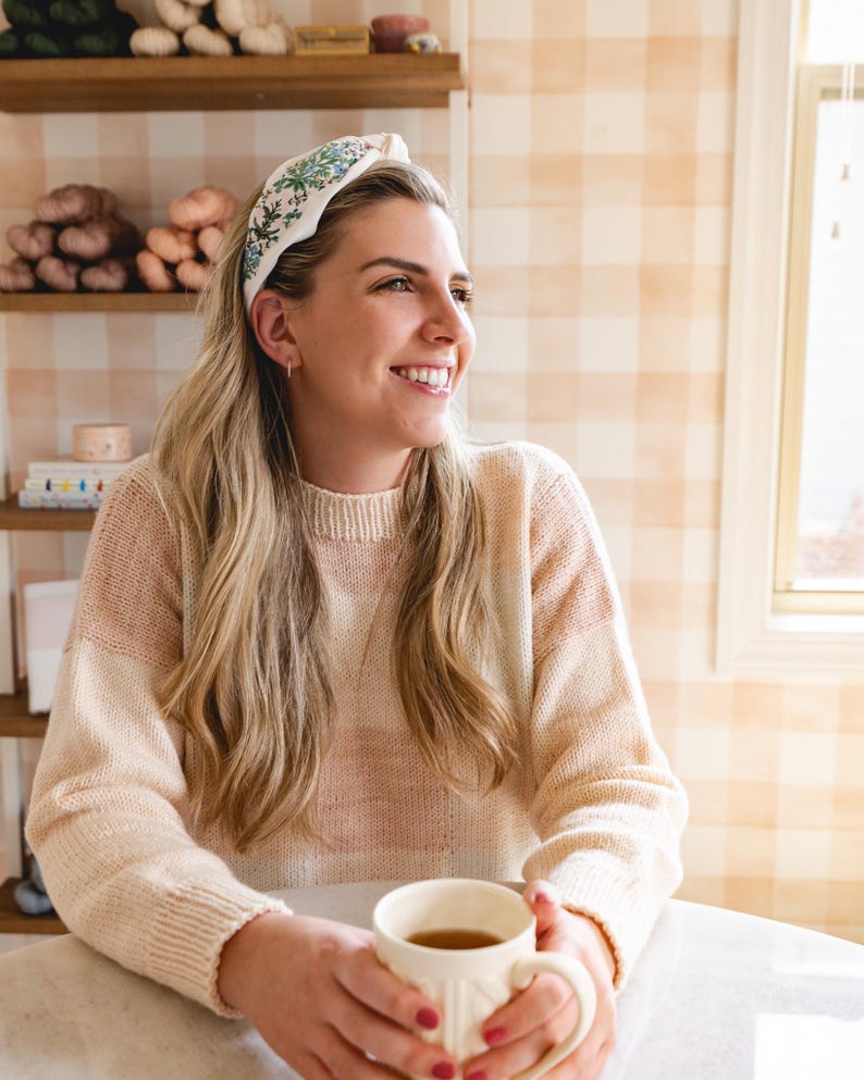 Op de afbeelding: Een cr&egrave;mekleurige hoofdband met een bloemenpatroon wordt gedragen door een persoon met lang blond haar. De persoon draagt een cr&egrave;me en roze trui en houdt een witte mok vast. De achtergrond heeft planken met garen en een geruite muur.