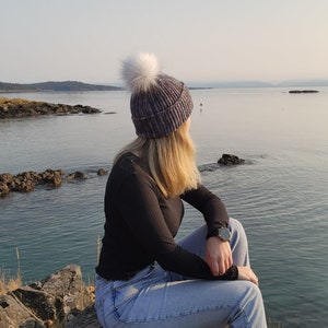 May include: A person wearing a blue and white knit beanie with a white pom pom sits on a rocky shore overlooking the ocean.