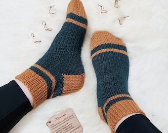 May include: A pair of teal and brown knit socks with a striped pattern. The socks are on a white fuzzy surface with a wooden tag that says "Kitchener" and has instructions for knitting.