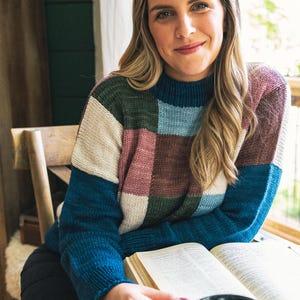 Op de afbeelding: Een kleurbloktrui in tinten blauw, groen, roze en cr&egrave;me. De trui heeft een hoge halslijn en wordt gedragen door een persoon die op een houten stoel zit. Een open boek en een donkergroene mok staan op de voorgrond.