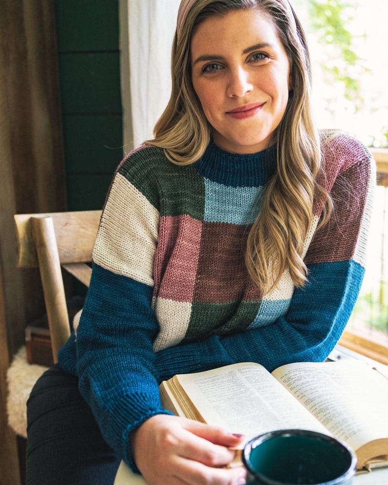Op de afbeelding: Een kleurbloktrui in tinten blauw, groen, roze en cr&egrave;me. De trui heeft een hoge halslijn en wordt gedragen door een persoon die op een houten stoel zit. Een open boek en een donkergroene mok staan op de voorgrond.