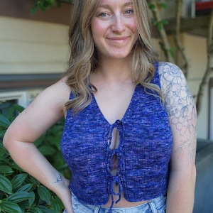 May include: A blue and purple knitted top with a lace-up front. The sleeveless top is worn with light wash denim jeans. The knitwear has a textured appearance and a V-neckline.