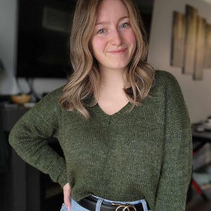 May include: A woman wearing a green knitted sweater with a v-neck and a black belt with a gold double ring buckle. She is wearing blue jeans.