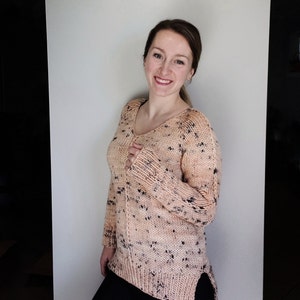 May include: A peach-colored sweater with black flecks throughout. The sweater has a v-neck and long sleeves. The sweater is knit and has a relaxed fit.