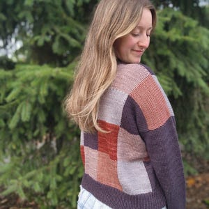 Op de afbeelding: Een patchwork trui in tinten paars, roest en grijs. De trui heeft een vierkant patroon op de rug en mouwen. De persoon draagt een lichtblauwe spijkerbroek.