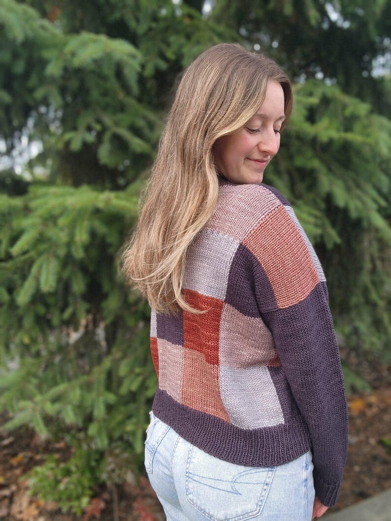 Op de afbeelding: Een patchwork trui in tinten paars, roest en grijs. De trui heeft een vierkant patroon op de rug en mouwen. De persoon draagt een lichtblauwe spijkerbroek.