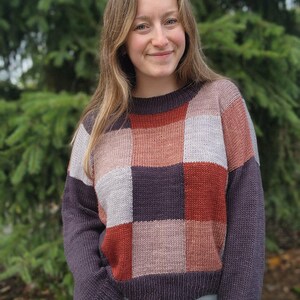 Op de afbeelding: Een patchwork trui in tinten paars, roest, roze en grijs. De trui heeft een ronde hals en lange mouwen. De persoon draagt een blauwe spijkerbroek. De achtergrond is een groene, onscherpe boom.