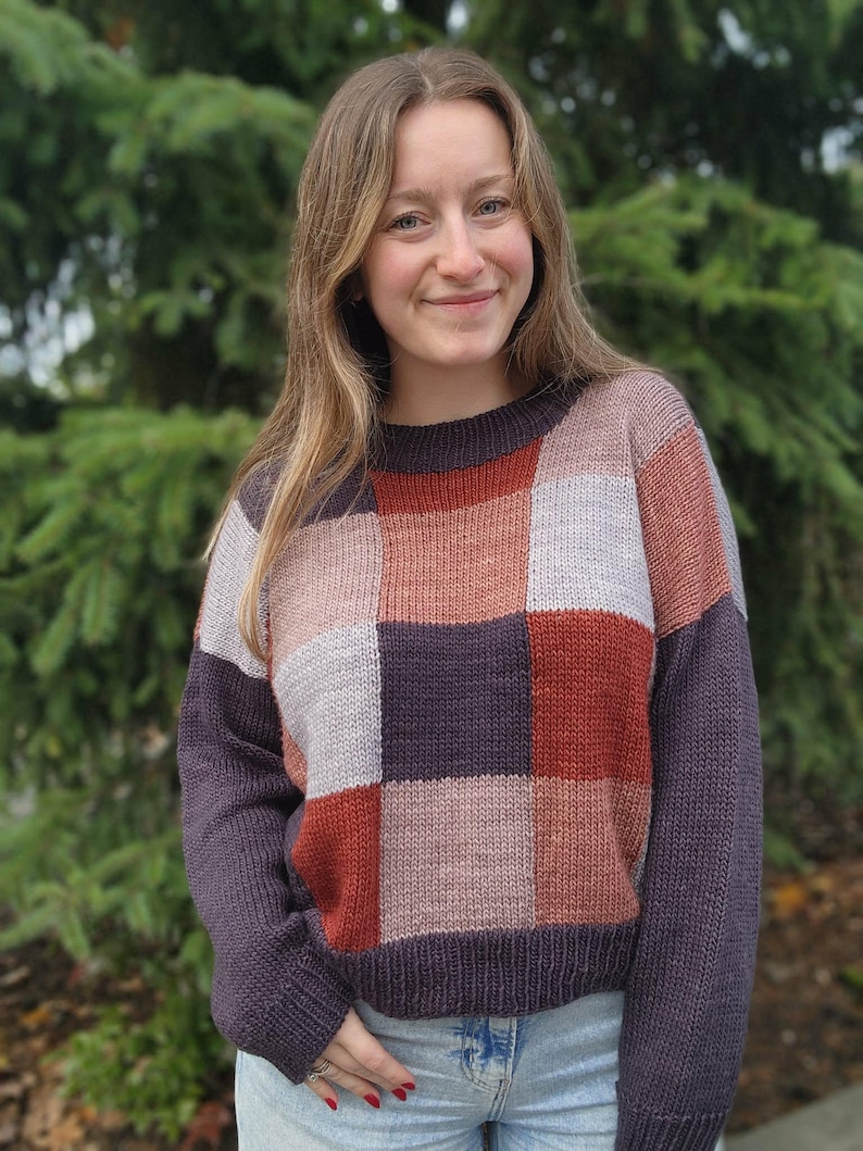 Op de afbeelding: Een patchwork trui in tinten paars, roest, roze en grijs. De trui heeft een ronde hals en lange mouwen. De persoon draagt een blauwe spijkerbroek. De achtergrond is een groene, onscherpe boom.