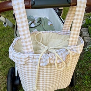 May include: A beige and white gingham patterned fabric bag with two long straps hanging from a stroller handle. The bag features a quilted exterior, a ruffled top edge, and a cream-colored rope drawstring closure to secure the contents inside.