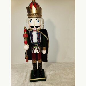 May include: A wooden nutcracker figurine with a red and gold crown, a green and black cape, and a white beard. The nutcracker is holding a red and gold staff.