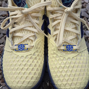 May include: Pair of cream-colored New Balance sneakers with blue and navy accents. The shoes feature a textured pattern on the toe box and a blue smiley face detail on the laces. The tongue has the New Balance logo.