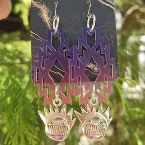 May include: A pair of dangle earrings with a geometric design in shades of purple and silver. The earrings feature a tribal-inspired pattern and a smiling face charm. The earrings are suspended from silver hooks.