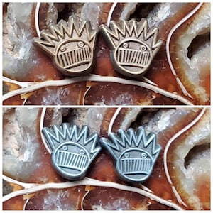 May include: Two pairs of earrings, one pair is gold and the other is silver. Both pairs feature a cartoon-like face with a crown of spikes.