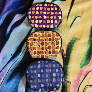 May include: Three oval-shaped zippered pouches with a cat print design. The top pouch is purple with white and orange cats. The middle pouch is yellow with brown, orange, and white cats. The bottom pouch is blue with gray cats.