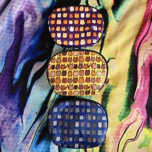May include: Three oval-shaped zippered pouches with different cat-themed patterns. The top pouch is purple with orange and white cats. The middle pouch is yellow with brown and orange cats. The bottom pouch is blue with gray cats.