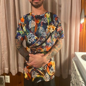 May include: A person wearing a black baseball cap, glasses, and a colorful t-shirt with a black strap. The t-shirt has a psychedelic pattern with a variety of colors, including red, orange, yellow, green, blue, and purple. The person is wearing a black watch on their left wrist and has tattoos on both arms.