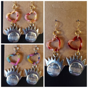 May include: Two pairs of earrings with a silver charm of a cartoon face with a crown of spikes. The top charm is a heart with a pink, orange, and white swirl pattern. The bottom charm is a red heart with a white swirl pattern.