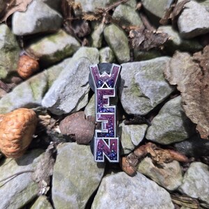 May include: A rectangular pin with the letters "WEHN" in a vertical arrangement. The letters are purple with a glittery, blue and purple fill. The pin has a silver border and is set against a background of gray rocks and leaves.