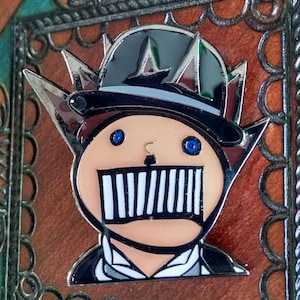 May include: A metal enamel pin featuring a stylized character wearing a black hat with silver spikes. The character has a light skin tone, bright blue eyes, and a wide mouth covered by vertical black and white bars, set against a dark background.