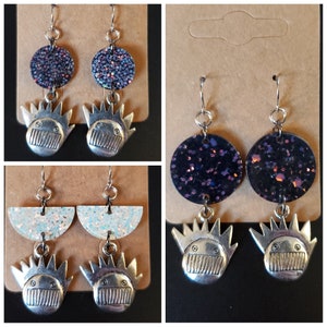 May include: Two pairs of dangle earrings with silver-toned charms. The first pair features a black and purple glitter circle with a silver-toned crown charm. The second pair features a white glitter half-circle with a silver-toned crown charm.