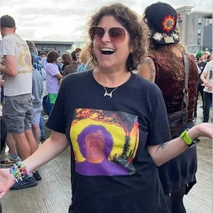 May include: A person wearing a black t-shirt with a colorful graphic of a man with purple hair and a yellow background. The person is wearing sunglasses and a silver necklace. A colorful bracelet is on the wrist.