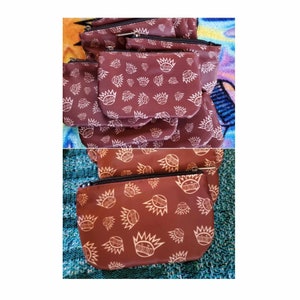 May include: A stack of brown zippered pouches with a white repeating pattern of a crown and a circle. The pouches are made of a soft, water-resistant fabric.