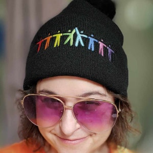 May include: Black knit beanie with a rainbow-colored chain of people holding hands embroidered on the front. The beanie has a black pom-pom on top.