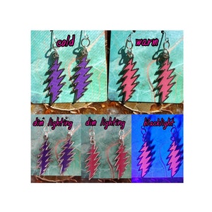 May include: A pair of silver earrings with a lightning bolt design. The earrings are shown in different lighting conditions, including cold, warm, dim lighting, and blacklight. The lightning bolt is purple in cold lighting, pink in warm lighting, and glows in the dark under blacklight.