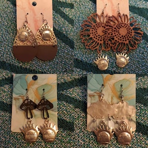 May include: Four pairs of earrings with different designs. The first pair is gold with a teardrop shape and a face design. The second pair is gold with a teardrop shape and a sunflower design. The third pair is black with a mushroom design and a face design. The fourth pair is silver with a cloud design and a face design.