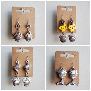 May include: Four pairs of silver earrings with different designs. The first pair features a flower charm and a skull charm. The second pair features a cheese charm and a skull charm. The third pair features a seashell charm and a skull charm. The fourth pair features a mermaid tail charm and a skull charm.