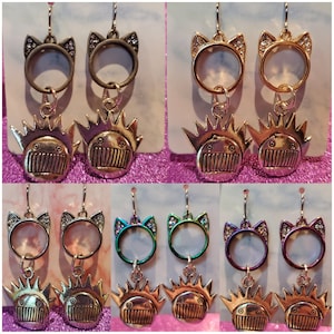 May include: A pair of earrings with a cat head charm and a cartoon face charm. The cat head charm is silver with rhinestones and the cartoon face charm is silver with a spiky crown. The earrings are available in three different colors: gold, green, and purple.