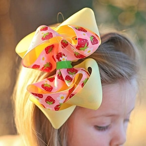 May include: A young girl with blonde hair wearing a large yellow and pink bow with a strawberry print. The bow is tied in a large, full knot.