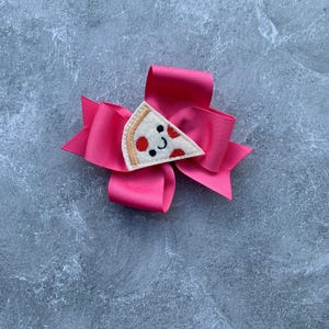 Puede incluir: Un lazo rosa para el pelo con una aplicaci&oacute;n de fieltro en forma de rebanada de pizza. La rebanada de pizza tiene pepperoni rojo y una cara sonriente.