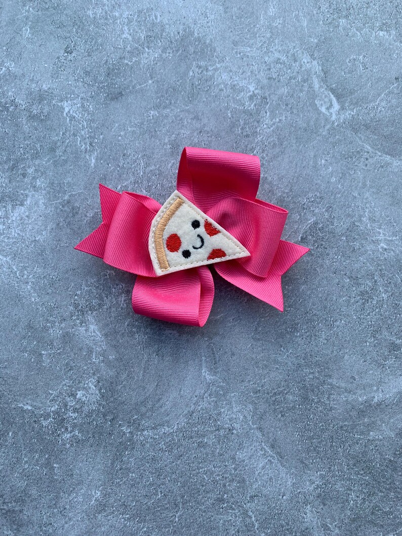 Puede incluir: Un lazo rosa para el pelo con una aplicaci&oacute;n de fieltro en forma de rebanada de pizza. La rebanada de pizza tiene pepperoni rojo y una cara sonriente.